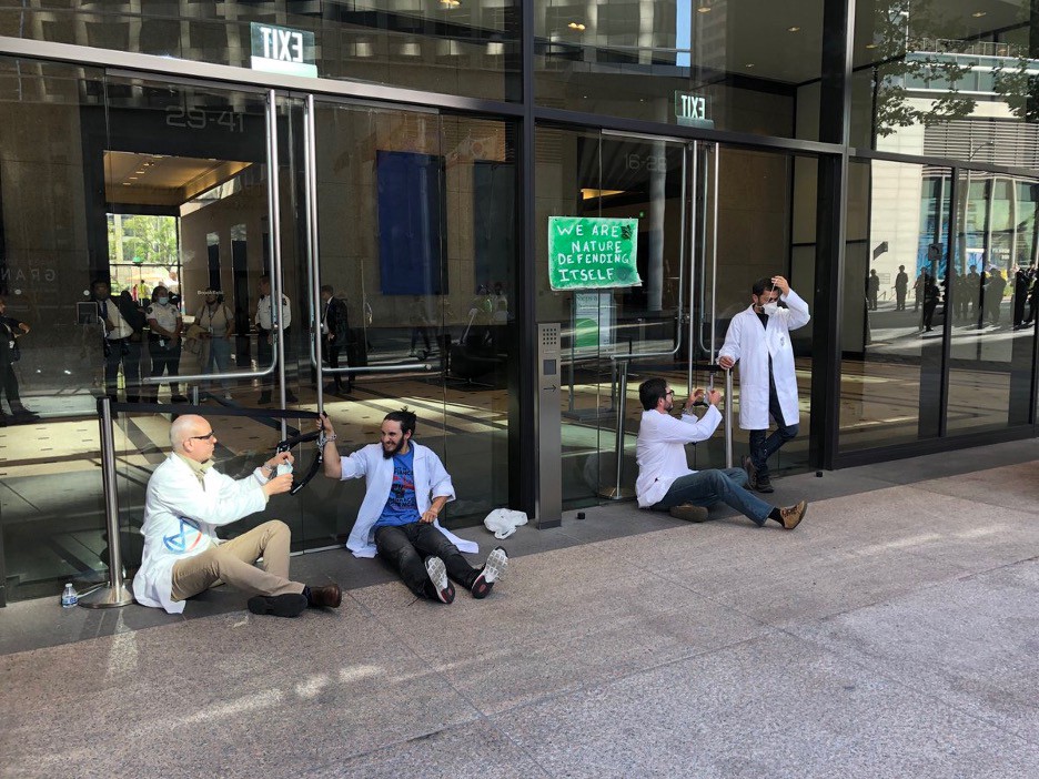 Image: Four scientists dressed in white lab coats sit on the ground, with their hands chained to large glass doors outside Chase bank