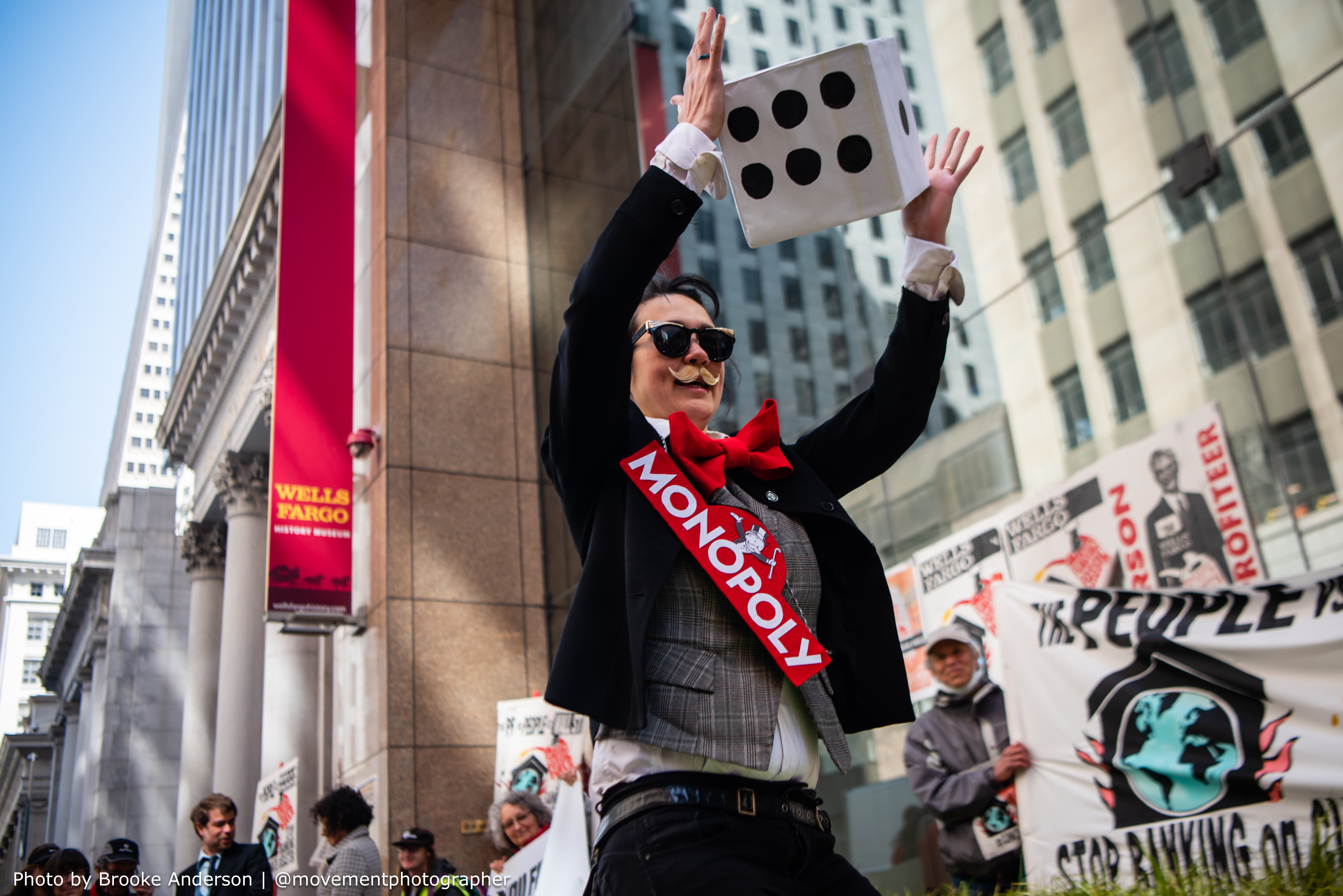 Character dressed in mustache and "Monopoly" sash holds a large cartoon die in front of Wells Fargo